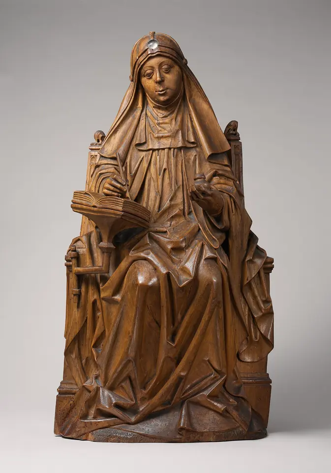 Carved wooden statuette of St. Bridget seated in a widow's veil and wimple, writing in a book held open on her knee