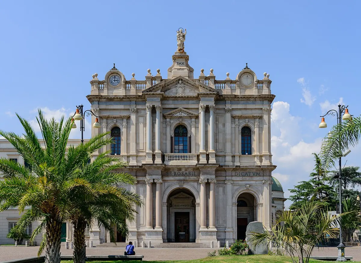 White marble facade of a large church with columns, arched doorways and a statue at the top, framed by palm and pine trees under a blue sky