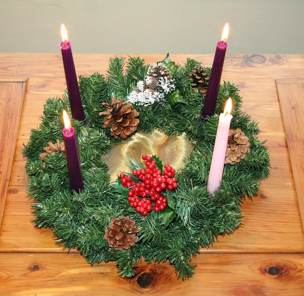 An evergreen wreath decorated with pine cones and red berries on a wooden table, holding three tall violet candles and one rose candle, all lit