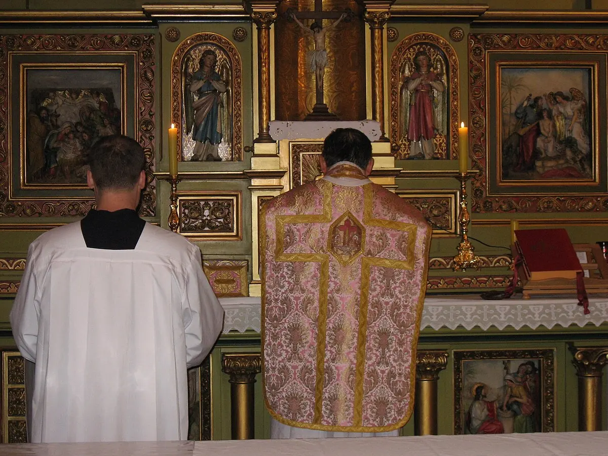 A priest seen from behind at an ornate wooden altar, wearing an embroidered rose-colored chasuble, with an altar server in a white surplice beside him