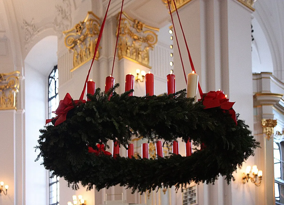 A huge evergreen wreath hung from red ribbons in a white and gold church, ringed with many red candles and a few tall white ones