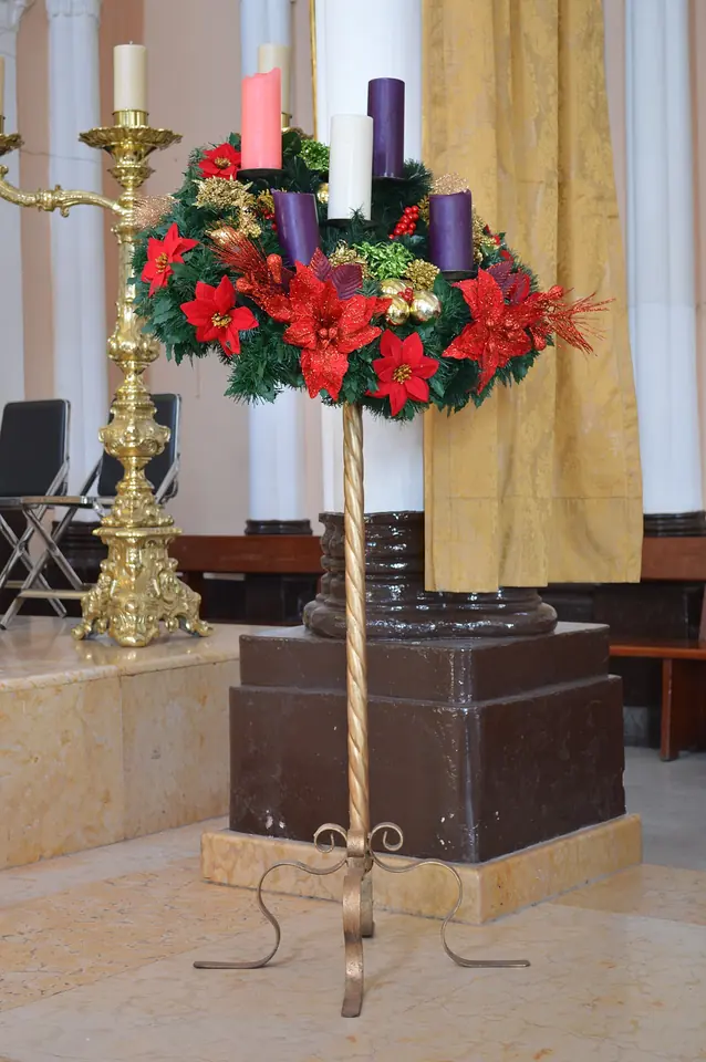 An Advent wreath of greenery and red poinsettias on a tall brass stand in a church, holding three violet candles, one rose candle and a white candle in the center