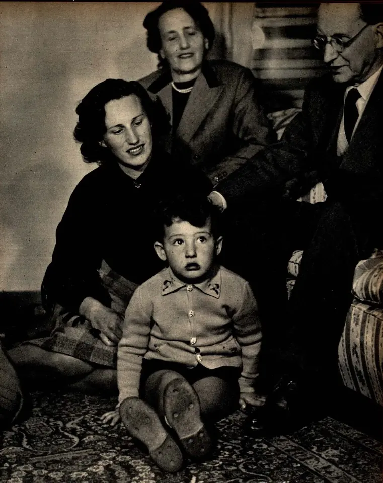 Black-and-white photograph of an older man with glasses sitting beside two women at home, with a small boy in a knitted sweater sitting on the rug in front of them