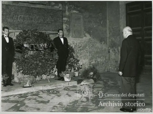 Black-and-white photograph of an elderly man in a dark suit standing with bowed head before a large wreath at a tomb, with two attendants beside it