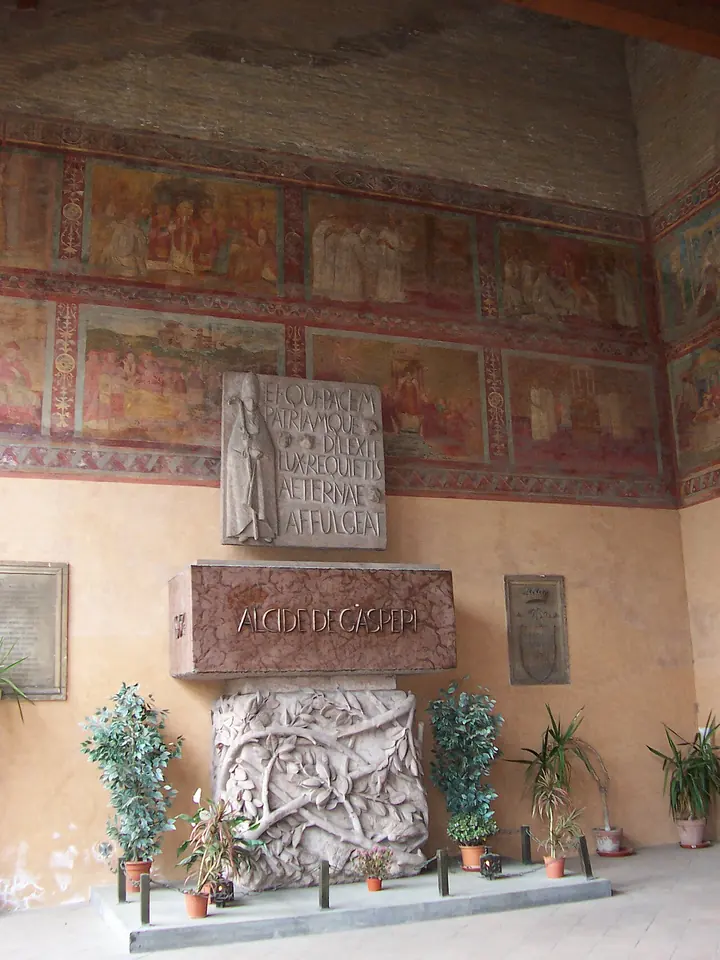 A stone tomb with carved reliefs and a Latin inscription against a frescoed wall in a church portico, with potted plants on either side