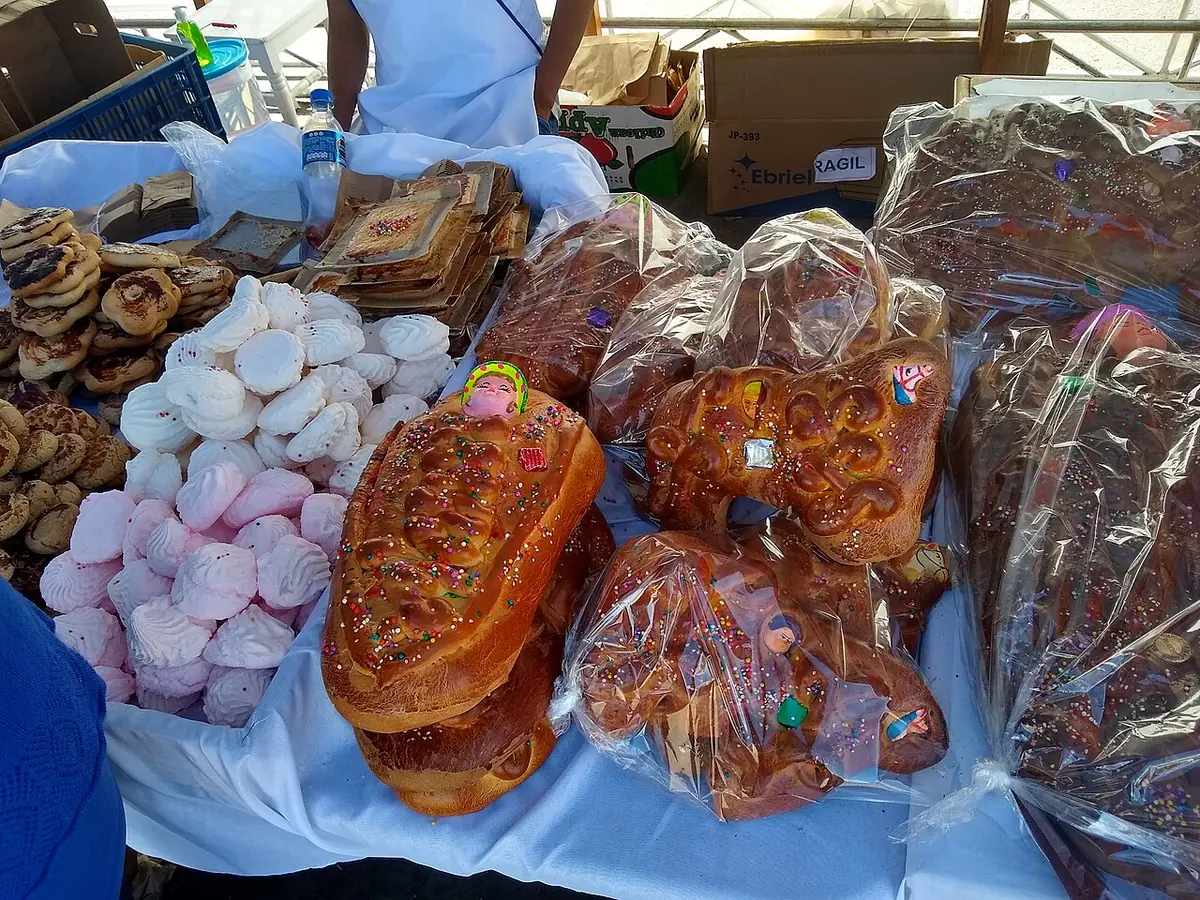 Market stall with large glazed loaves of sweet bread wrapped in plastic, some shaped like swaddled babies, next to piles of pink and white meringues
