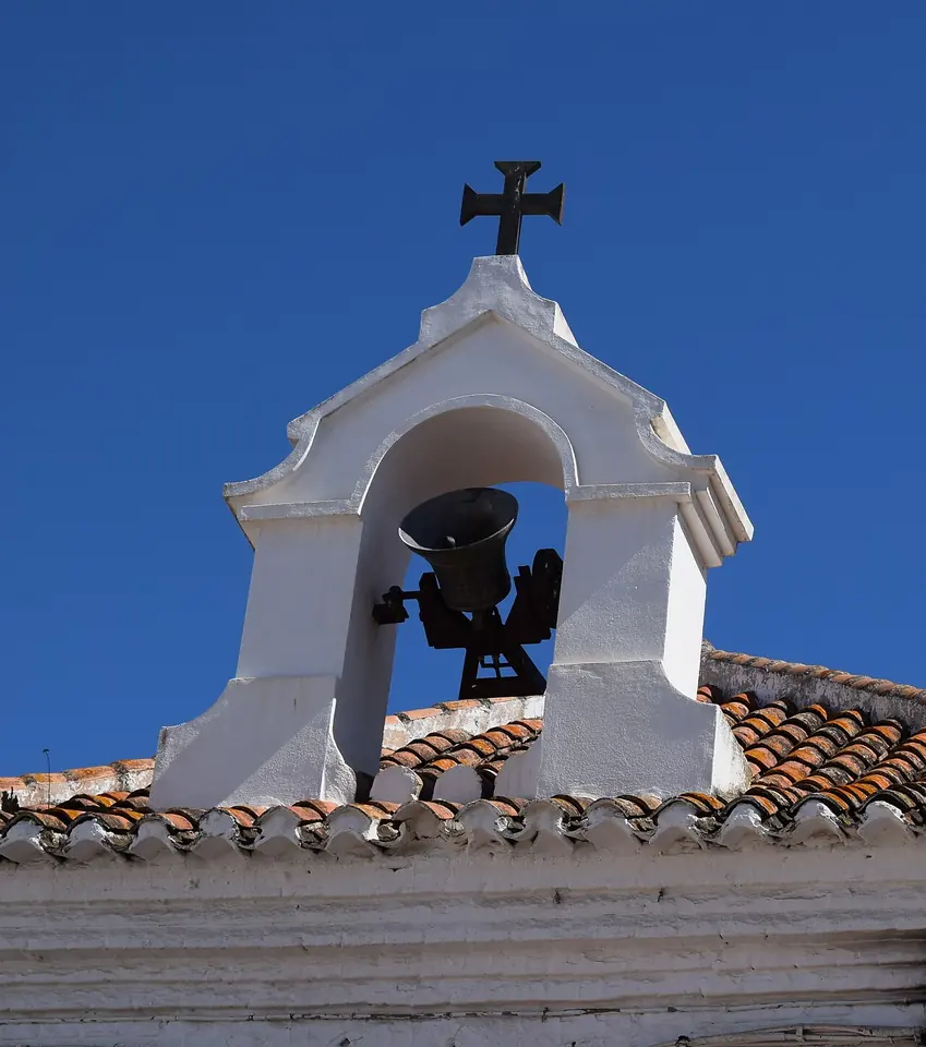 A small white bell gable with an iron cross on top and a dark bell swinging inside it against a deep blue sky