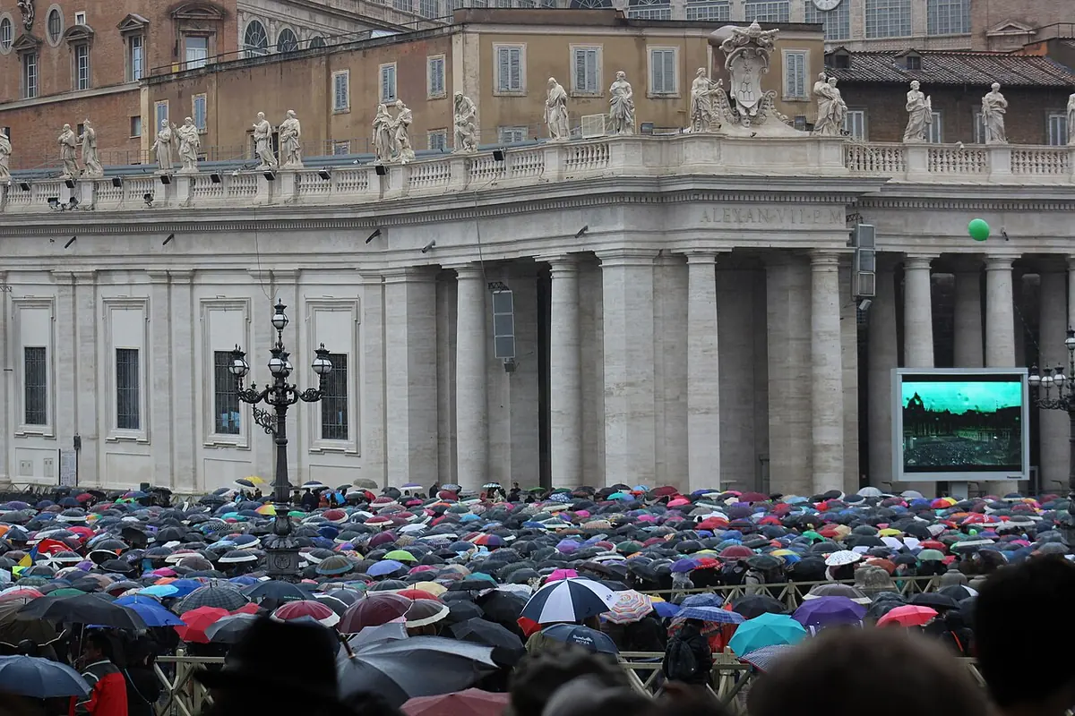 A large crowd with many umbrellas fills a square in front of a curved colonnade, with a big video screen showing the Pope