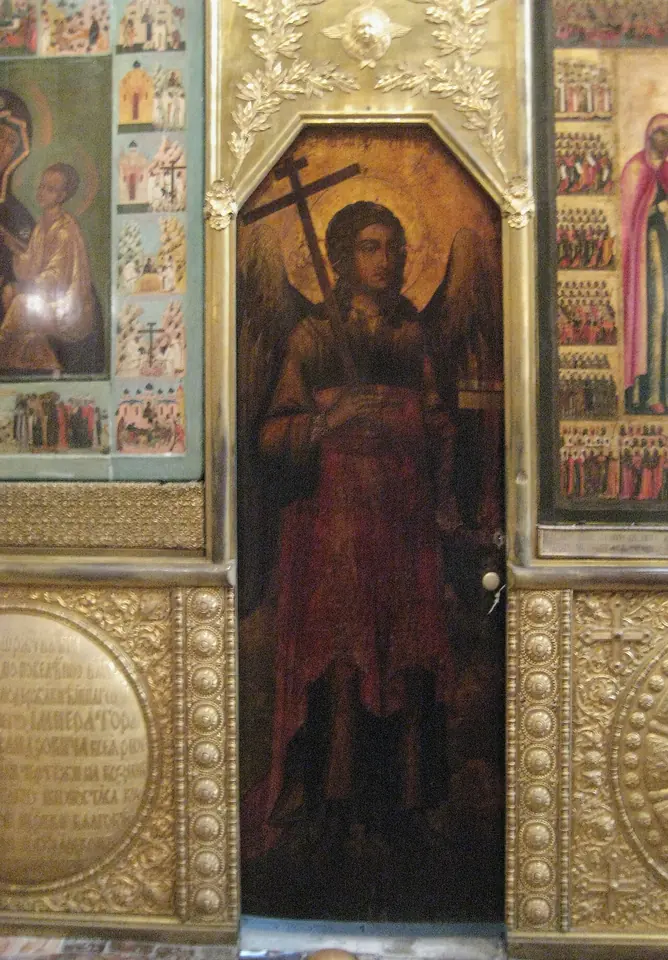 Dark old icon of a winged angel holding a tall cross, painted on a narrow door set into a gilded screen of icons