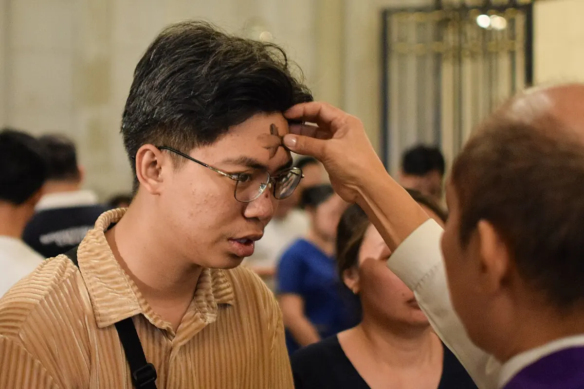 A young man in glasses closes his eyes as a minister's thumb traces a cross of ashes on his forehead during a crowded Ash Wednesday Mass