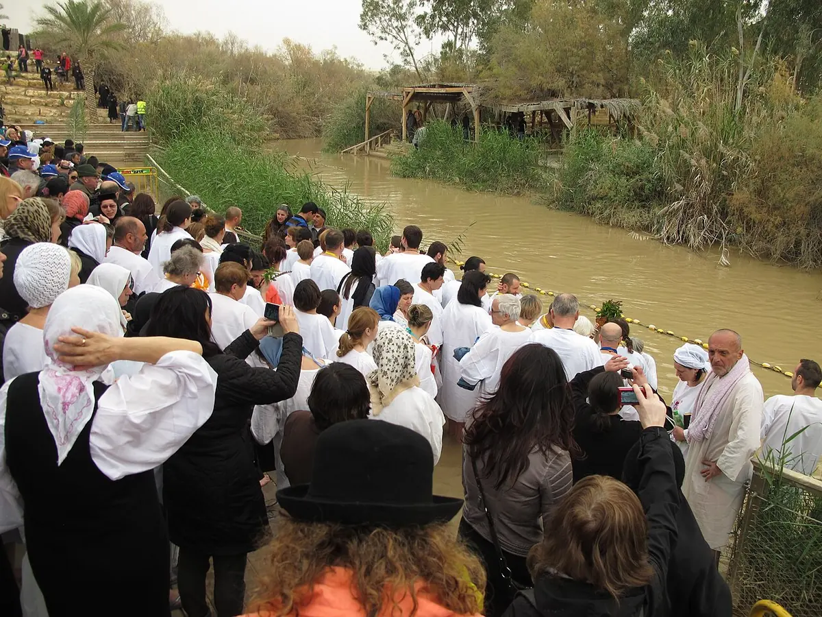 Crowds of pilgrims, many in white robes and head coverings, gathered on steps leading down to the muddy green Jordan River at the traditional baptism site