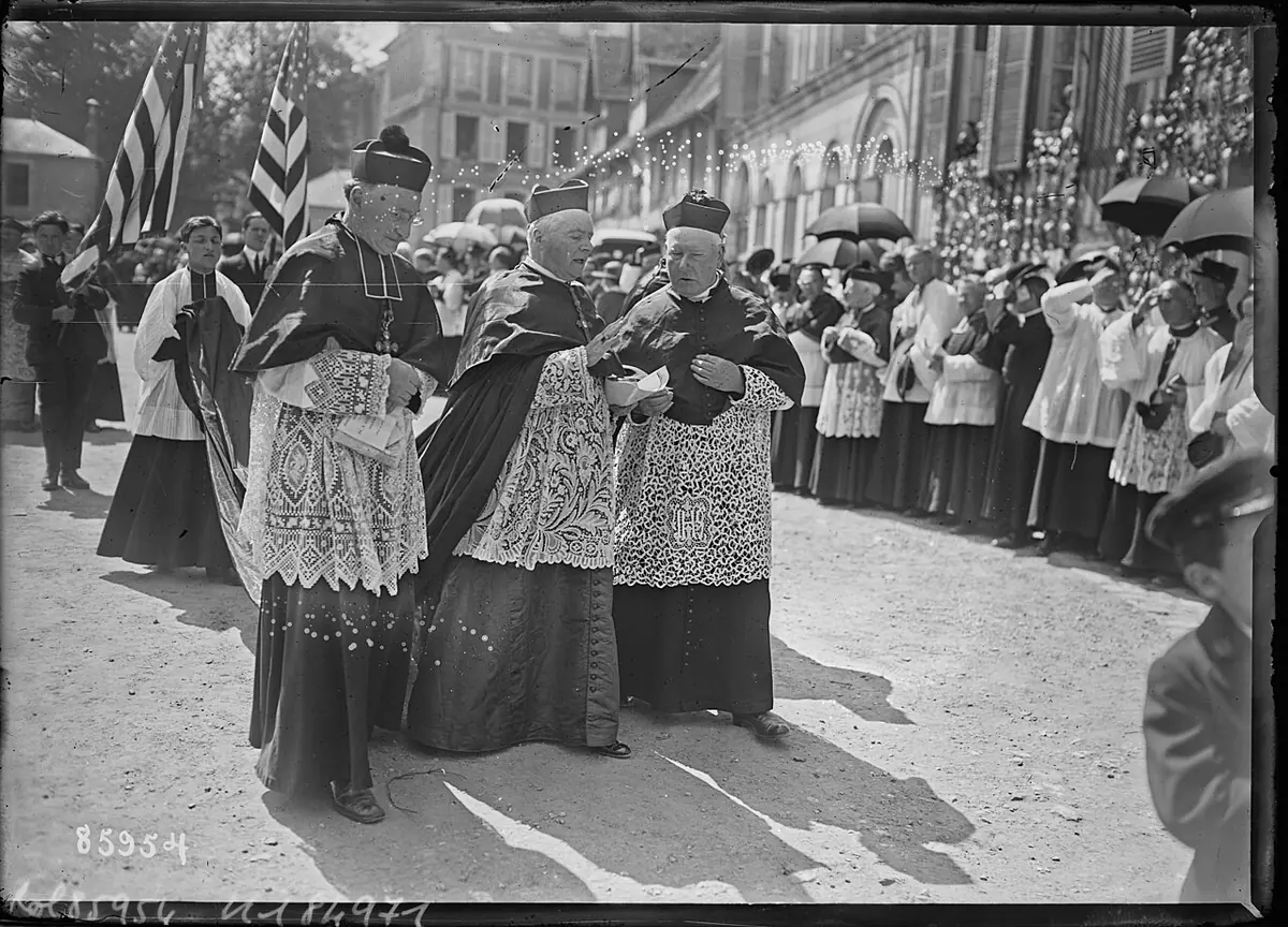 Black-and-white press photograph of a bishop in lace surplice and cape walking among clergy through a crowd in a street hung with flags