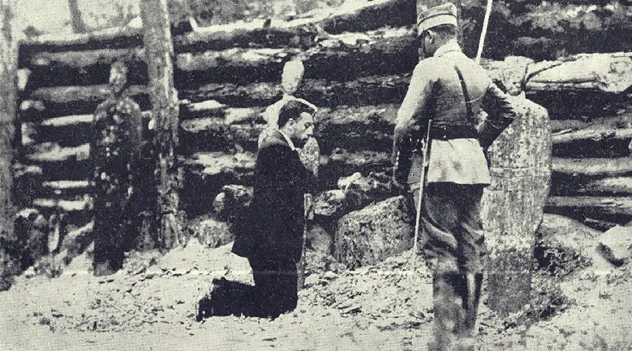 Grainy black-and-white news photograph of a man in a dark suit kneeling in prayer before a wooden wall, with uniformed soldiers standing nearby