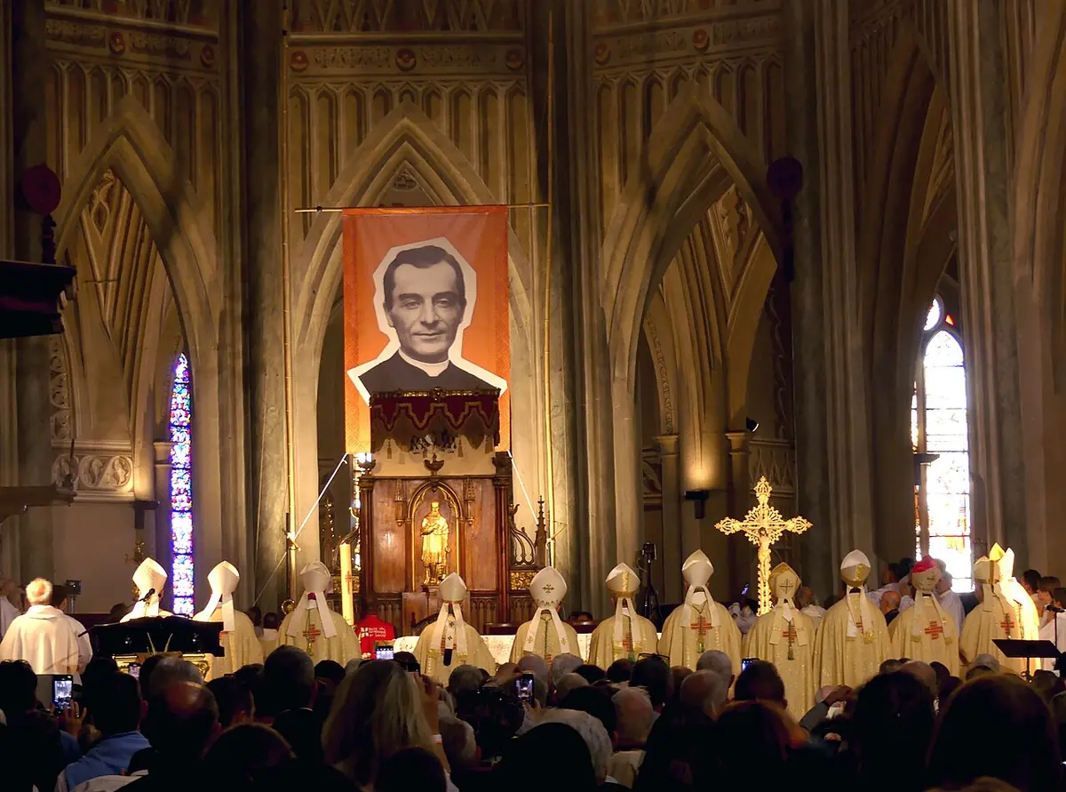 A large banner portrait of a smiling young priest hangs above the sanctuary of a Gothic cathedral, with a row of bishops in white miters and a crowded congregation below