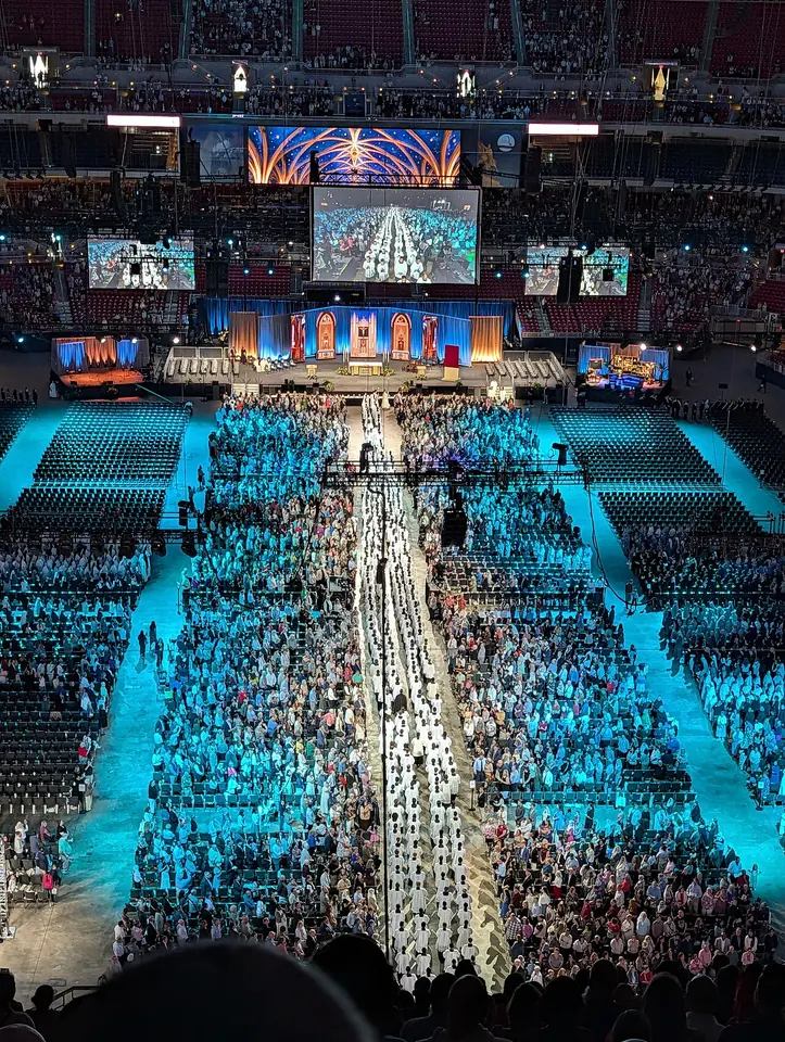 A vast indoor stadium filled with thousands of people facing a distant altar platform under large video screens, with a long center aisle lined with clergy in white