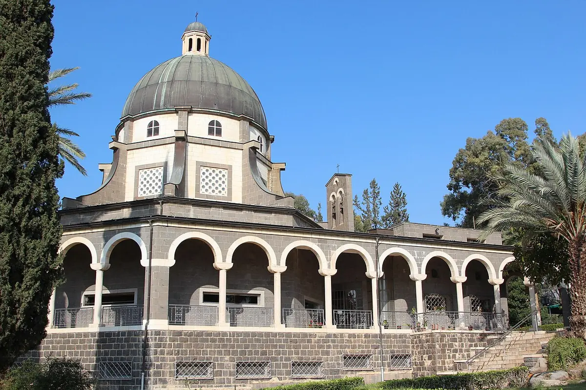 An eight-sided church of dark basalt with a white arcade around it and a gray dome, among palm trees and gardens under a blue sky