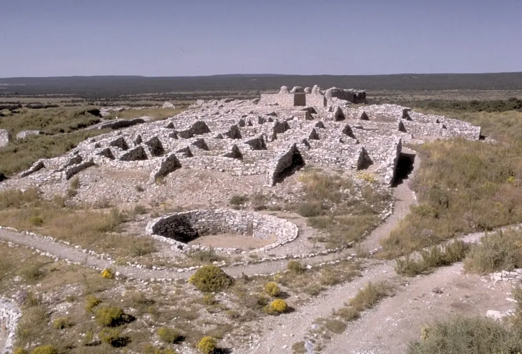 The low stone ruins of a pueblo and its round kiva on a dry, grassy plain in central New Mexico, under a pale sky