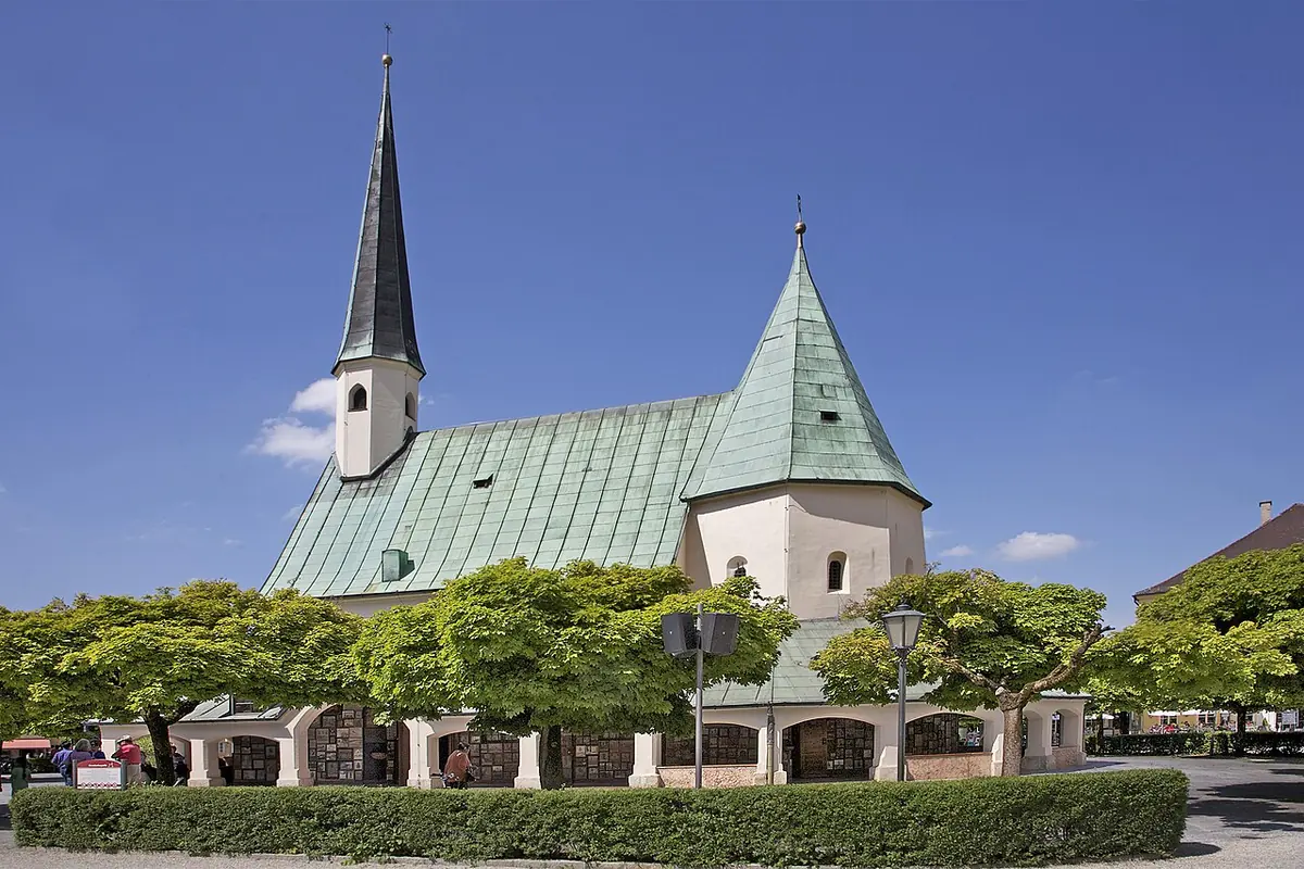 A small white chapel with a steep green roof and a tall slender spire, ringed by a covered walkway and trimmed trees on a sunny square