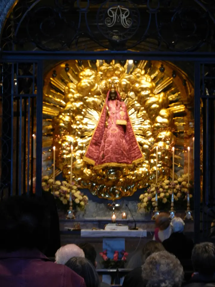 A dark-faced crowned statue of Mary and the Child dressed in a triangular red and gold robe, set in a golden sunburst above an altar, with worshipers in front