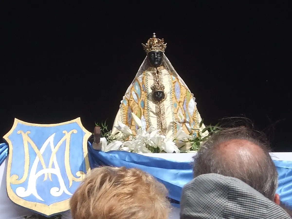The dark-faced crowned statue in a white and blue robe, surrounded by lilies, carried at night above the heads of the crowd beside a blue banner with the monogram of Mary