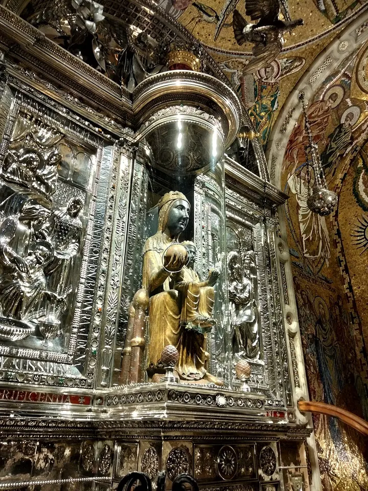 Gilded seated statue of the Virgin with a dark face holding the Child on her lap, framed by an ornate silver altarpiece with carved reliefs