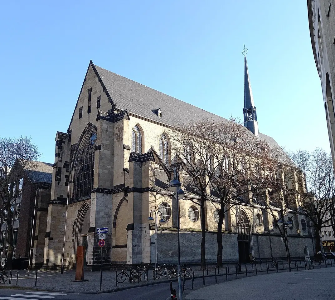 The long Gothic stone nave of the Minoritenkirche in Cologne with a slender spire, seen from the street under a blue sky