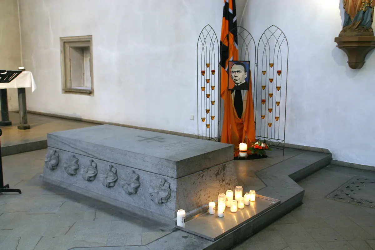 A plain gray stone sarcophagus on the floor of a white church chapel, with candles burning at its foot and a portrait of Kolping on a stand behind it