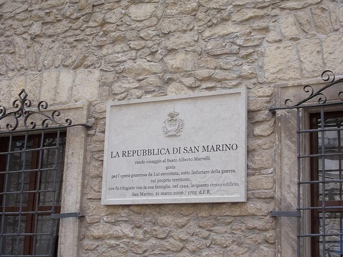 A white marble plaque with the arms of San Marino and an inscription honoring Blessed Alberto Marvelli, set in a stone wall between two barred windows