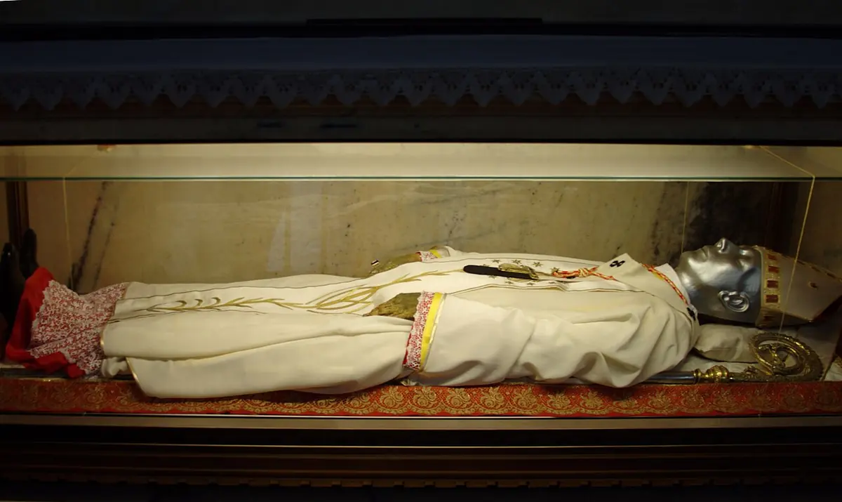 The body of a bishop in white vestments and miter, his face covered by a silver mask, lying in a glass case in a dark marble chapel