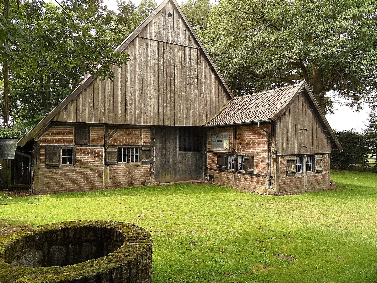 A low Westphalian farmhouse of brick and dark weathered planks with a steep gable roof, set on a lawn under tall trees, with an old stone well in front