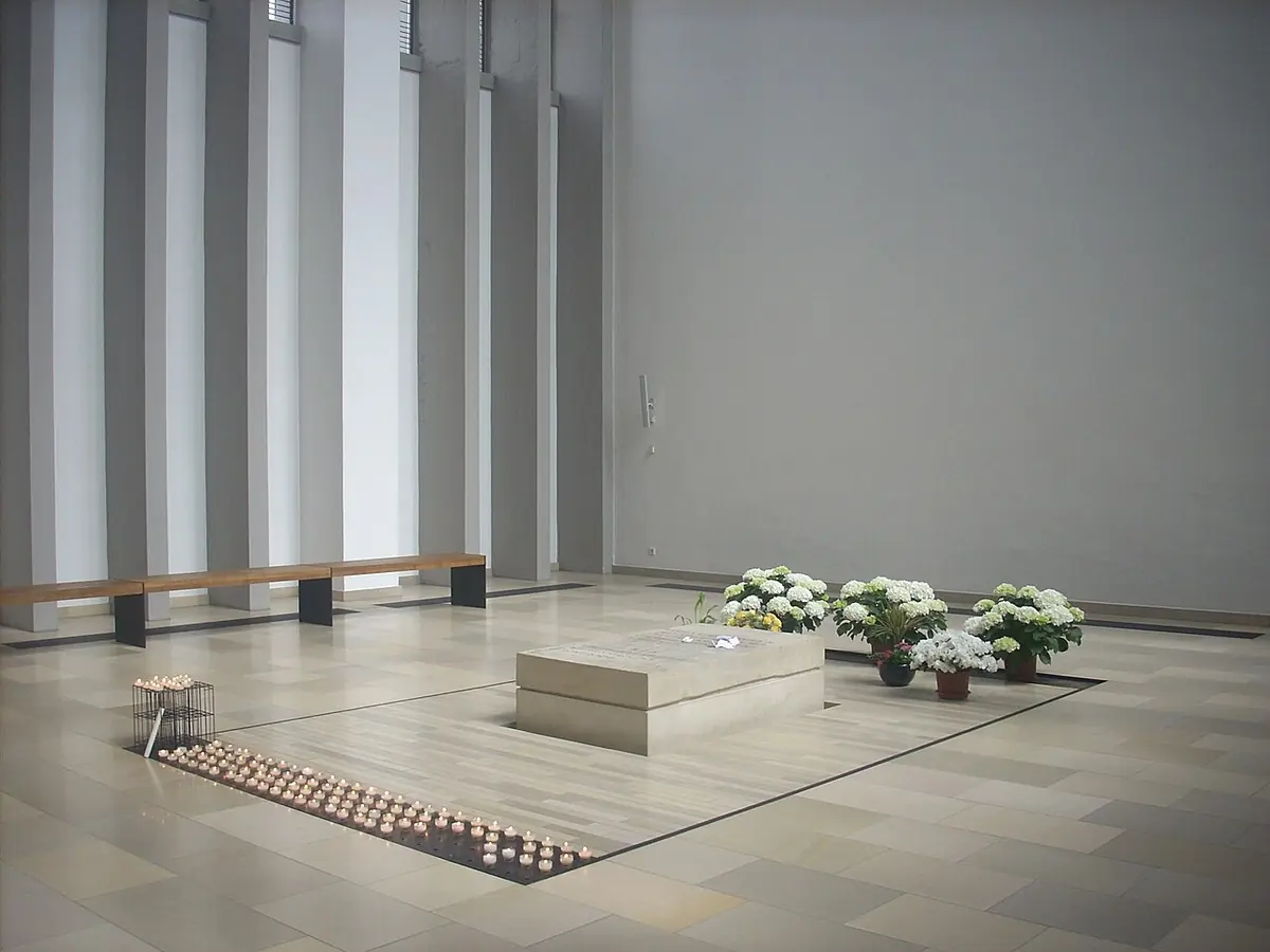 A plain stone tomb in a bright modern crypt with white walls, flowers on the floor beside it and rows of small votive candles in front