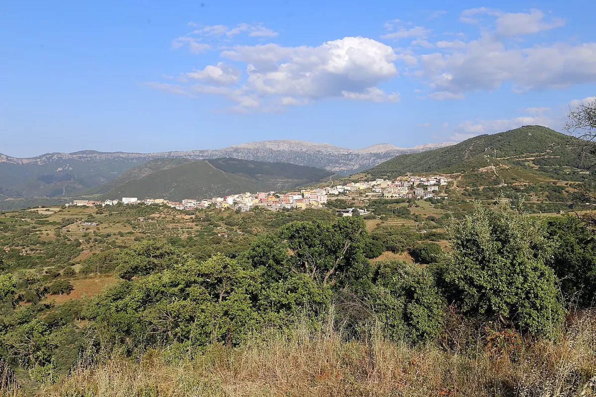 A distant view of a hill village of pale houses on a ridge, surrounded by green scrub and wooded mountains under a blue sky