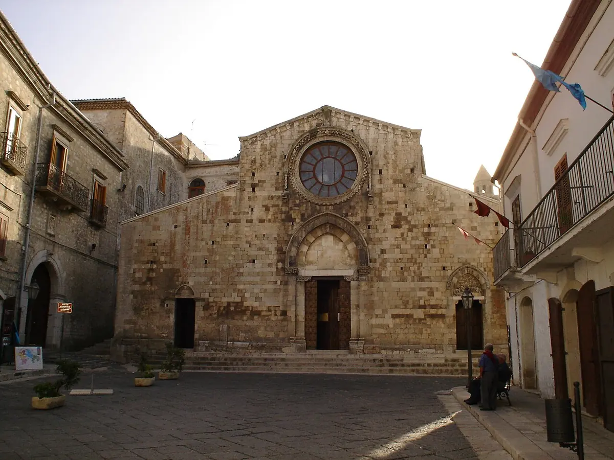 The plain Romanesque stone facade of Bovino Cathedral with a round rose window over the door, on a paved square between old houses