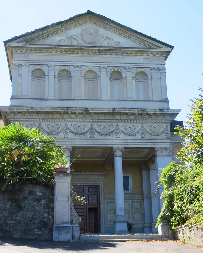 The neoclassical facade of the Stresa sanctuary with columns, a pediment and arched windows, framed by palms and green trees