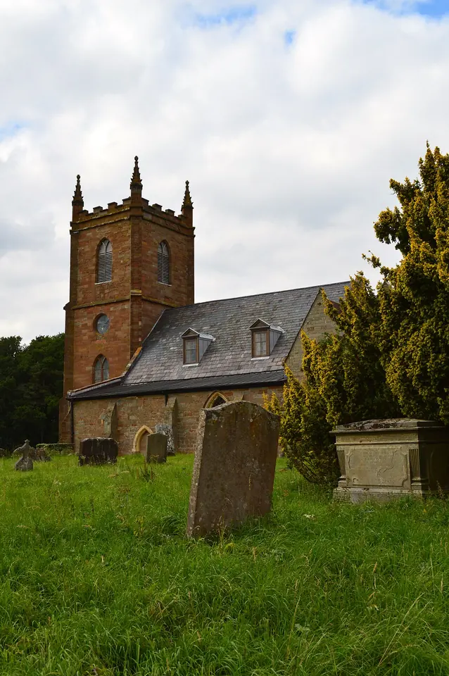 Old English parish church of reddish stone with a square battlemented west tower, seen across a grassy churchyard with leaning gravestones and a dark yew tree