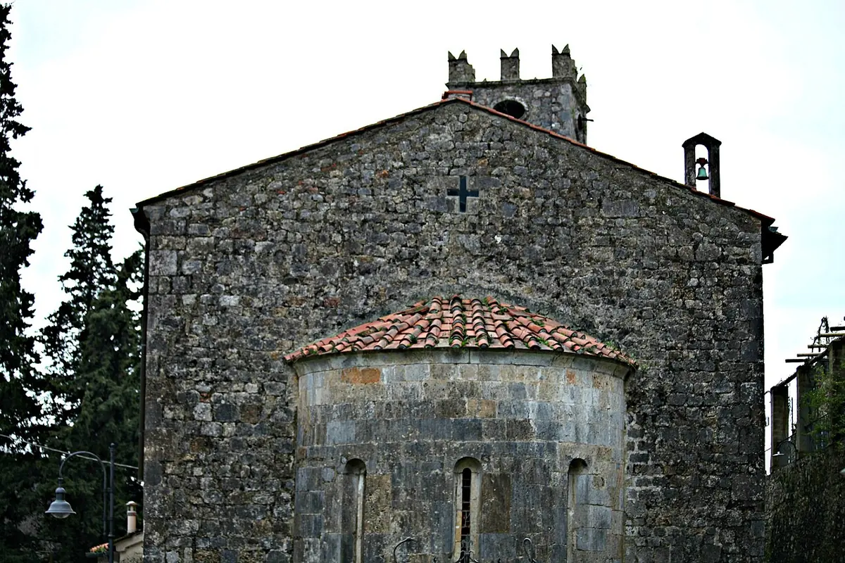 Small old church of rough gray stone with a rounded apse, a tiled roof, a cross on the gable and a little bell tower among cypress trees