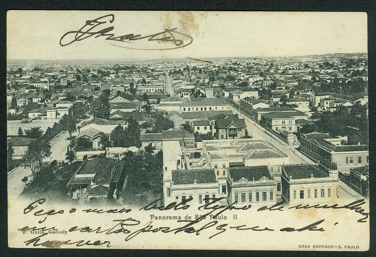 Old black-and-white postcard showing a bird's-eye view over the low tiled roofs and straight streets of São Paulo around 1900, with handwriting across the top and bottom
