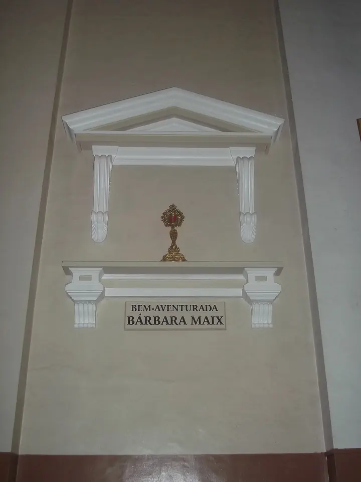 Small golden reliquary on a white wall shelf under a carved pediment, above a plaque reading Bem-Aventurada Bárbara Maix