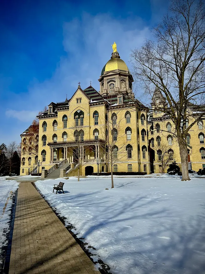 The yellow-brick Main Building of the University of Notre Dame with its gold dome and statue of Mary, under a blue sky with snow on the lawn