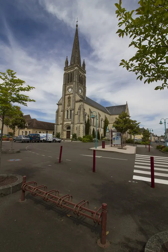 Village church of pale stone with a tall pointed spire on a square, with a crosswalk and young trees in the foreground