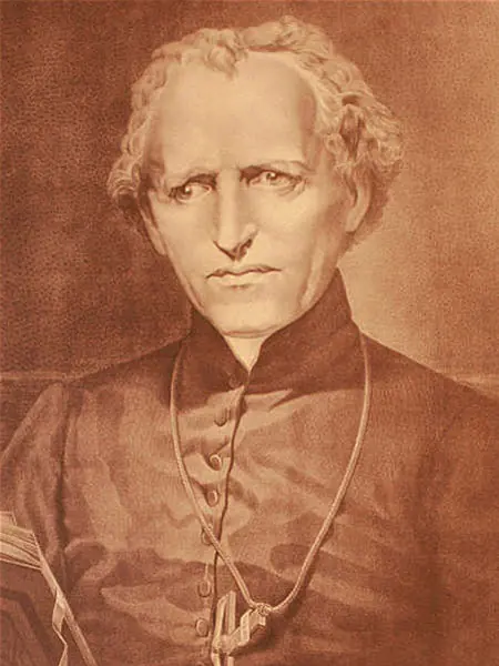 Sepia portrait of a priest in his sixties with long wavy hair and a serious expression, in a black cassock with a cross on a cord