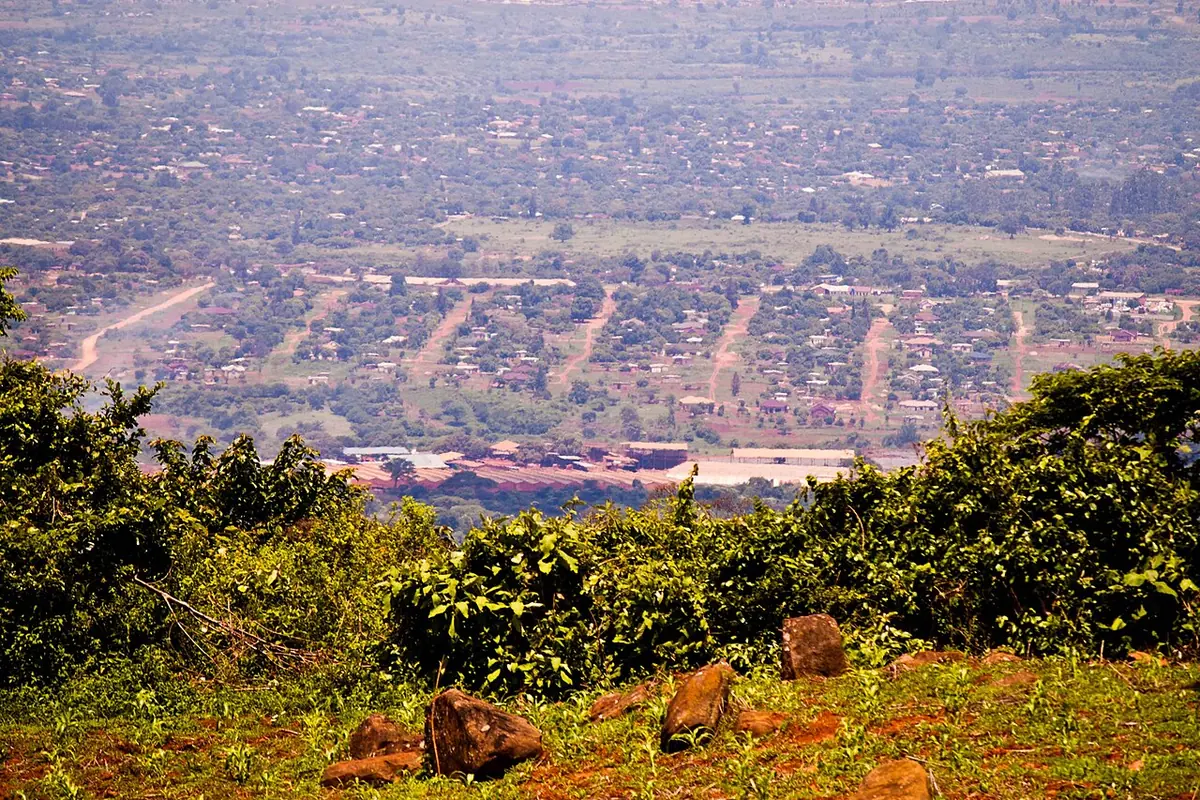 A wide view over a town of small houses and streets spread across a plain, seen from a hillside with red rocks and green bushes