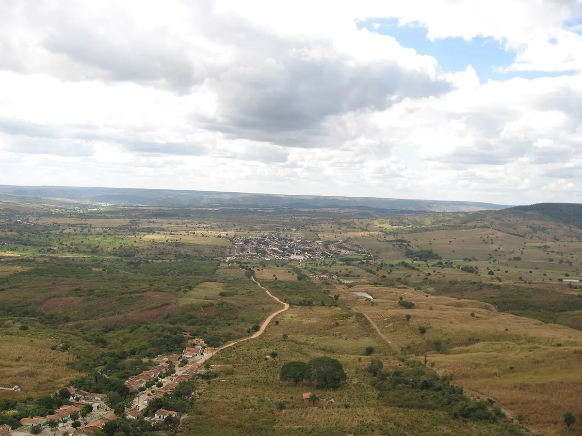 A wide view from a height over a small town of white houses and a church tower, set among green and brown fields and scrub, with low hills on the horizon