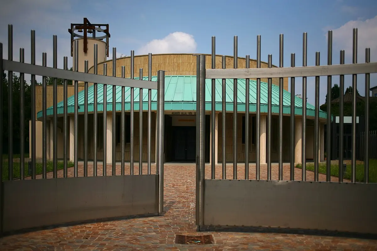 A round modern church with a green copper roof and a tall cross, seen through an open gate of vertical metal bars