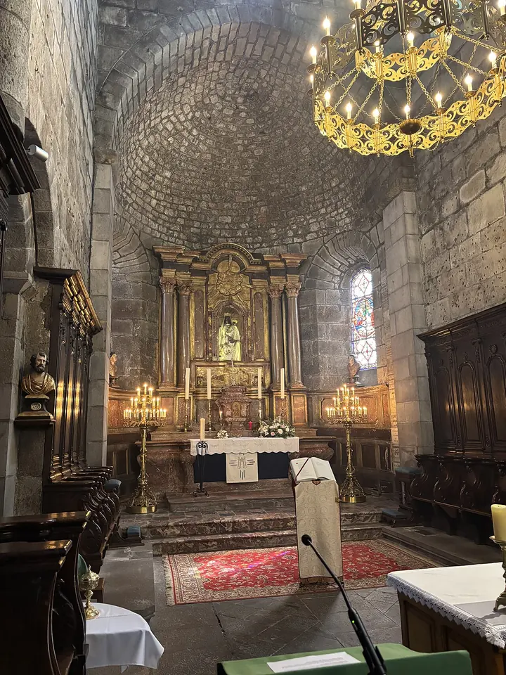 Inside a Romanesque stone church, a gilded altarpiece and altar beneath a stone half-dome, with a brass chandelier in the foreground