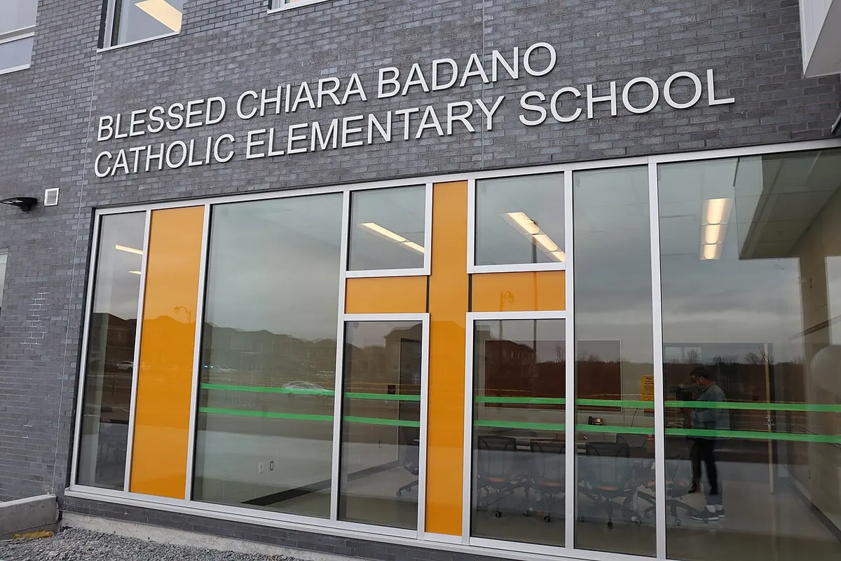 The gray brick front of a modern school with large windows, an orange cross worked into the glass and the name Blessed Chiara Badano Catholic Elementary School
