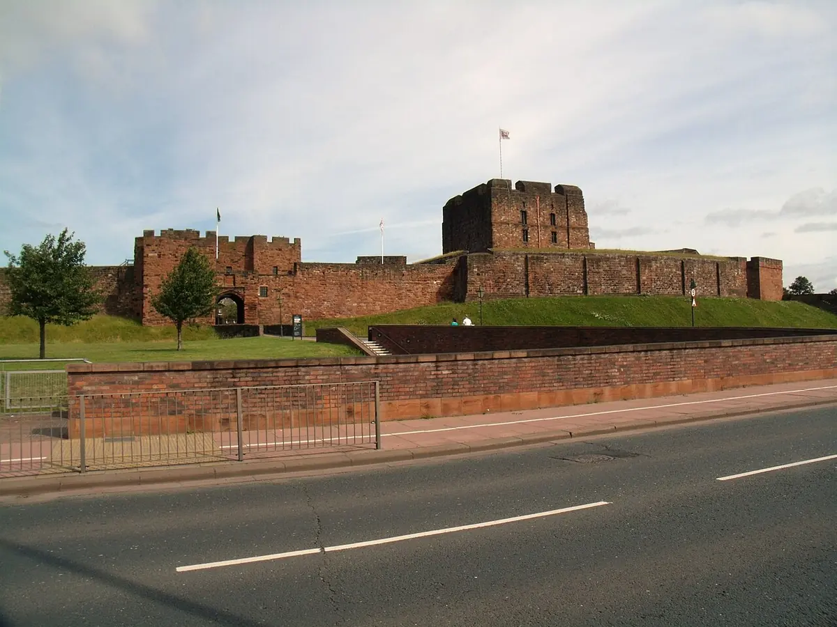 The red sandstone walls and square keep of Carlisle Castle above a grassy bank and a road