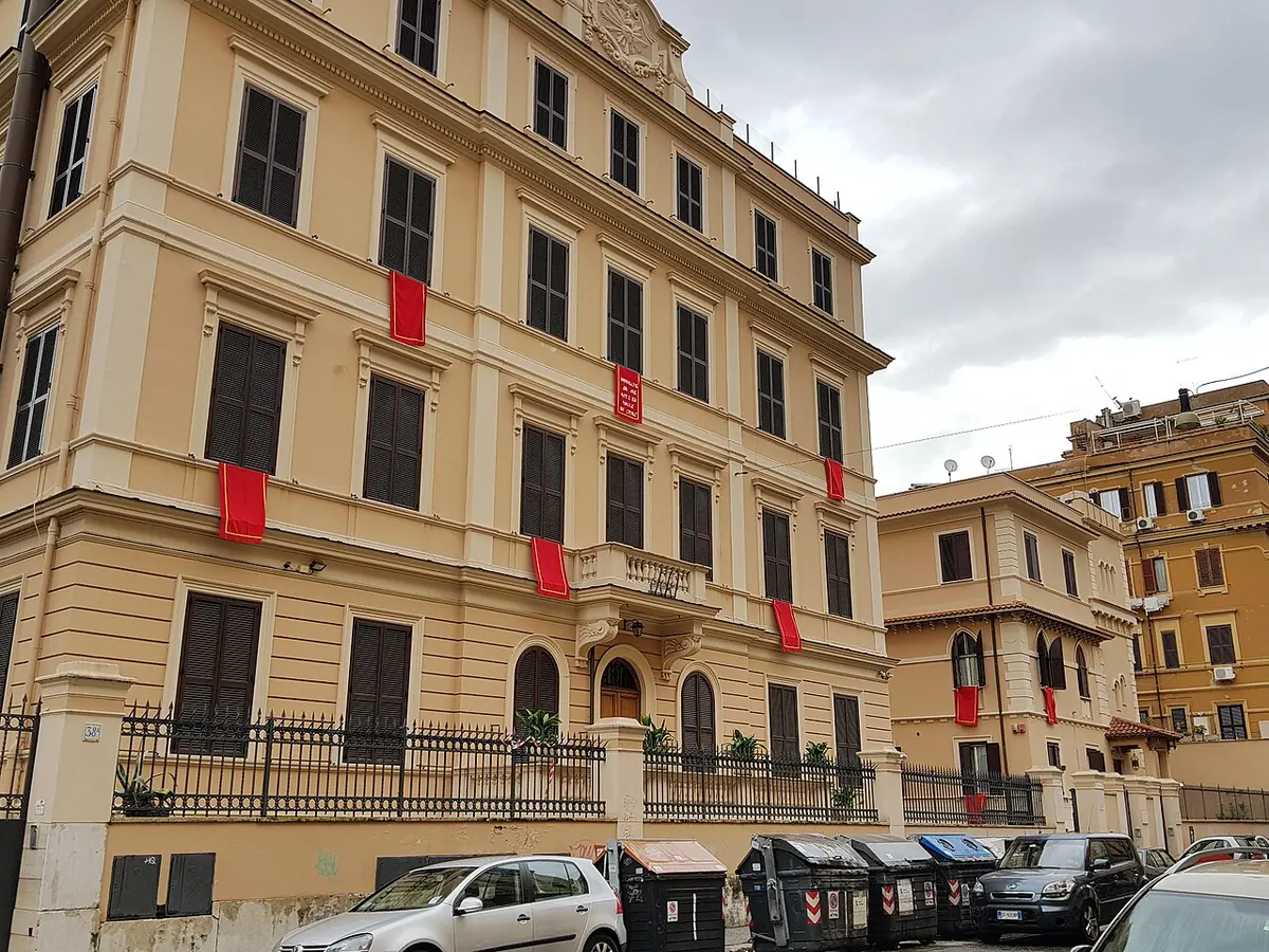 A pale yellow five-story building on a Roman street, with red banners hanging from the windows