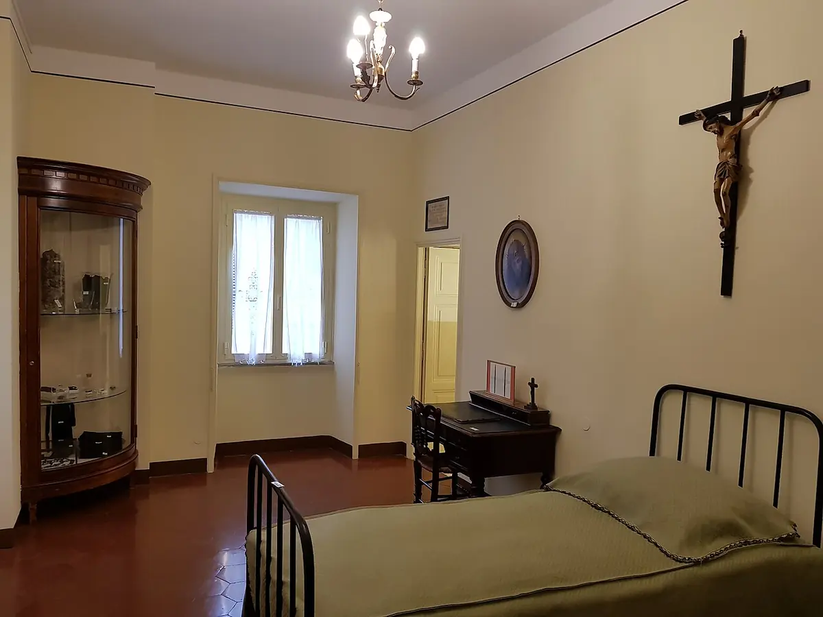 A simple room with an iron bed, a small desk, a glass cabinet and a crucifix on the wall