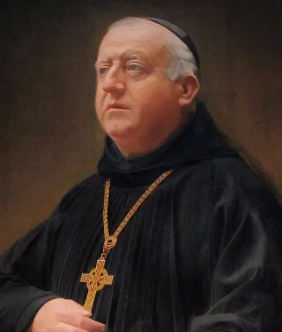 Photograph of a heavy-set older monk in a black habit and skullcap wearing a large cross on a chain, looking up to the left